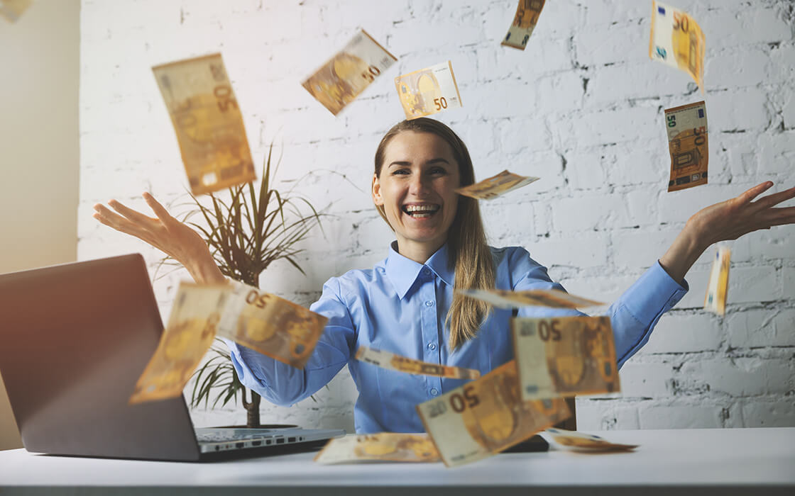 Bestbezahlte Berufe: Eine junge Frau sitzt am Arbeitsplatz im Büro, vor ihr fliegen Geldscheine